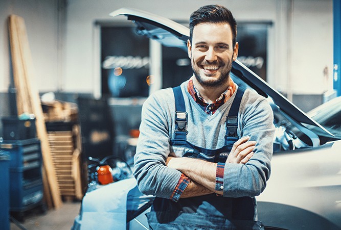 mechanic-standing-in-front-of-car A mechanic smiling with his arms folded standing in front of the side of a vehicle with its hood up.